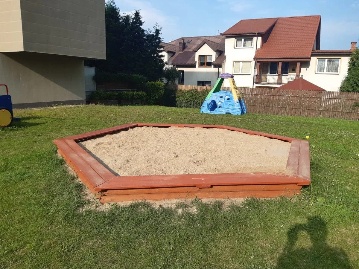 Drewniana piaskownica na trawniku przy domach, wypełniona świeżym piaskiem